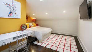 Une chambre avec une lucarne, un lit agrémenté de coussins colorés, un bureau avec une chaise, un téléviseur fixé au mur et un tapis à carreaux rouges et blancs.