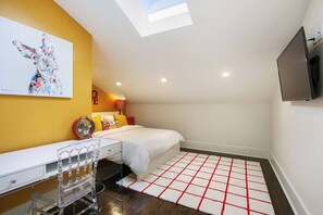 A bedroom with a skylight, a bed with colorful pillows, a desk with a chair, a TV on the wall, and a red and white checkered rug.
