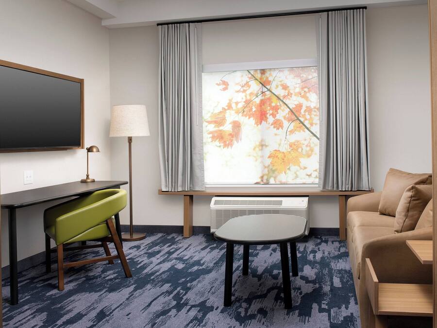 A hotel room with a desk, a green chair, a sofa, a window with curtains, and a radiator.