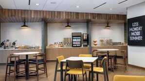 Een modern café met houten details, een centraal gelegen toonbank met een koffiemachine en zithoeken met gele stoelen.
