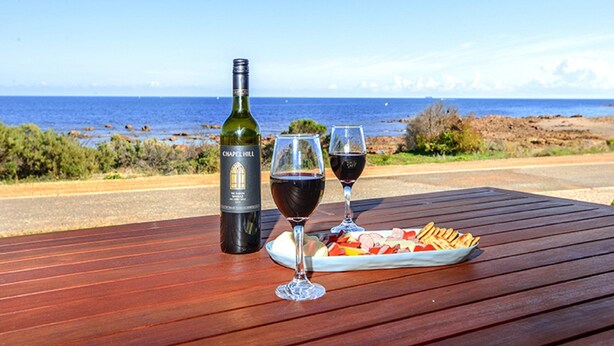 Une table en bois, une bouteille de vin, deux verres à vin et une planche de charcuterie, le tout installé sur une route en bord de mer.
