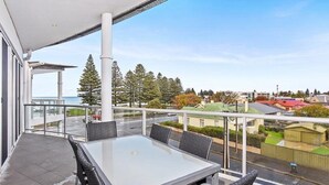 A balcony with a glass railing, a table, and chairs. - 3BR Penthouse w/ Ocean Views – Pool, Spa Bath, Tennis Court & Secured Parking | Ideal for Extended Stays (Victor Harbor)