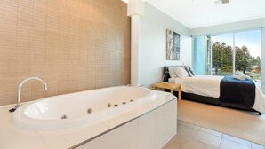A modern bathroom with a large bathtub and a separate bedroom with a bed and a view of trees. - 3BR Penthouse w/ Ocean Views – Pool, Spa Bath, Tennis Court & Secured Parking | Ideal for Extended Stays (Victor Harbor)