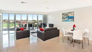 A modern living room with a large glass door leading to a balcony, a dining area with white chairs, and a painting on the wall. - 3BR Penthouse w/ Ocean Views – Pool, Spa Bath, Tennis Court & Secured Parking | Ideal for Extended Stays (Victor Harbor)