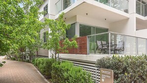 Een modern gebouw met een glazen balkon, een verharde looppad en een bord dat de ingang aangeeft.