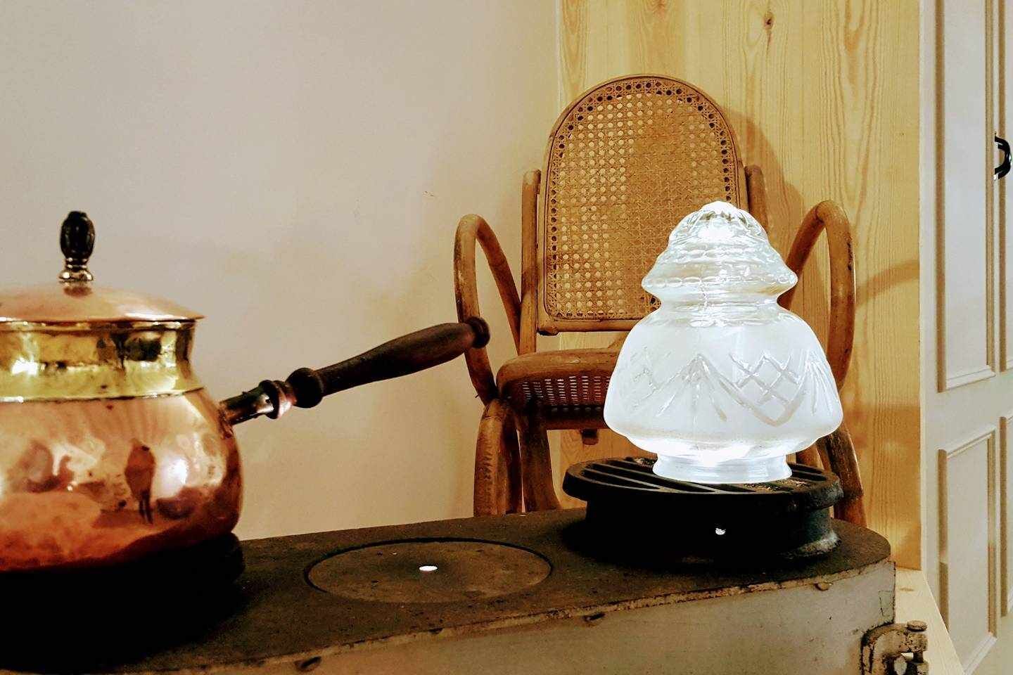 A brass kettle on a stove, a glass vase on a stand, and a wicker chair in the background.