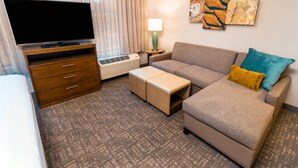 Habitación de hotel con televisor de pantalla plana, muebles de madera, un sofá con cojines, una lámpara y una obra de arte en la pared.