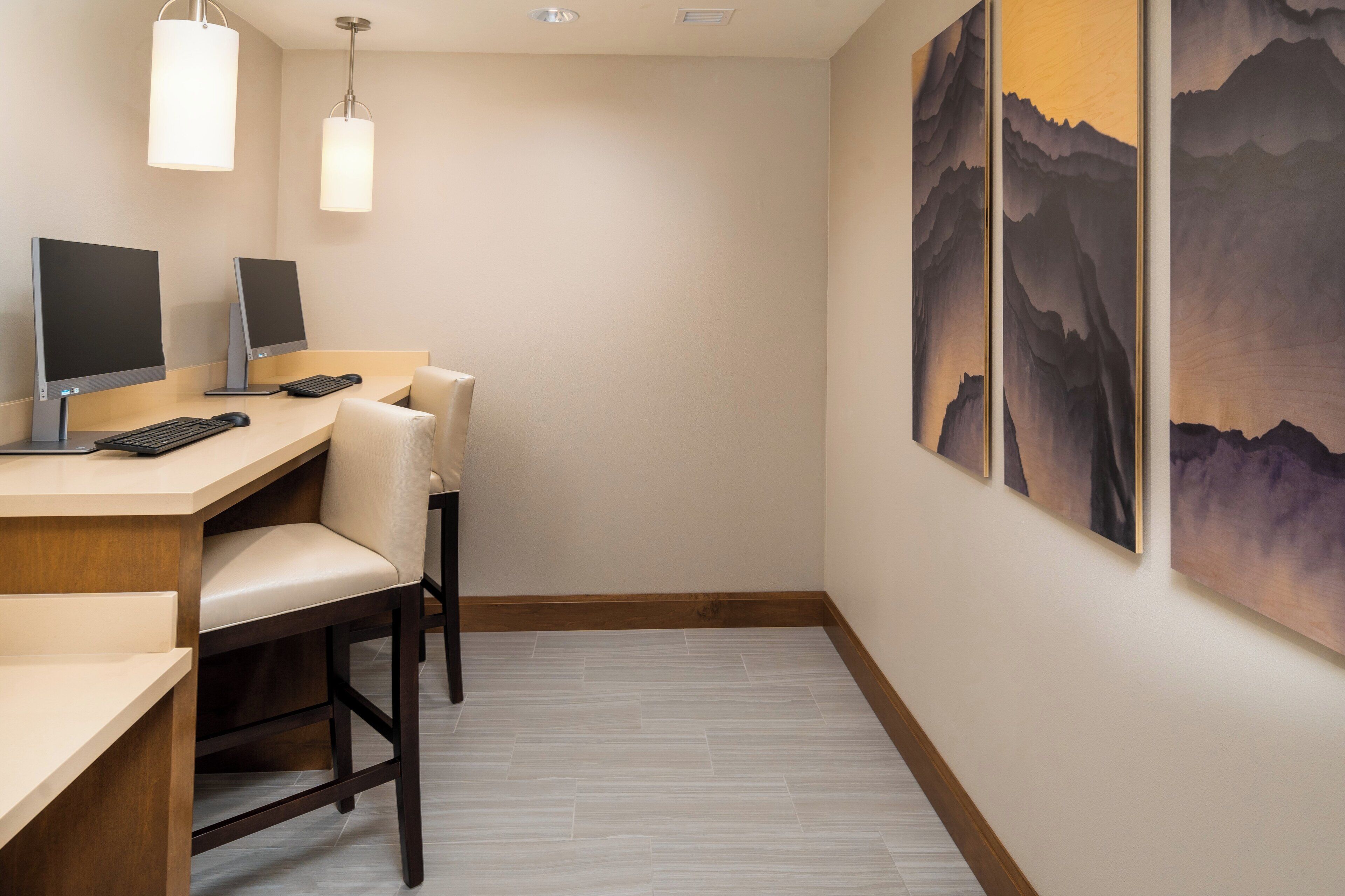 Un espace de bureau moderne comprenant un bureau, un ordinateur et deux chaises. On y trouve deux lampes fixées au mur et un tableau représentant des montagnes.