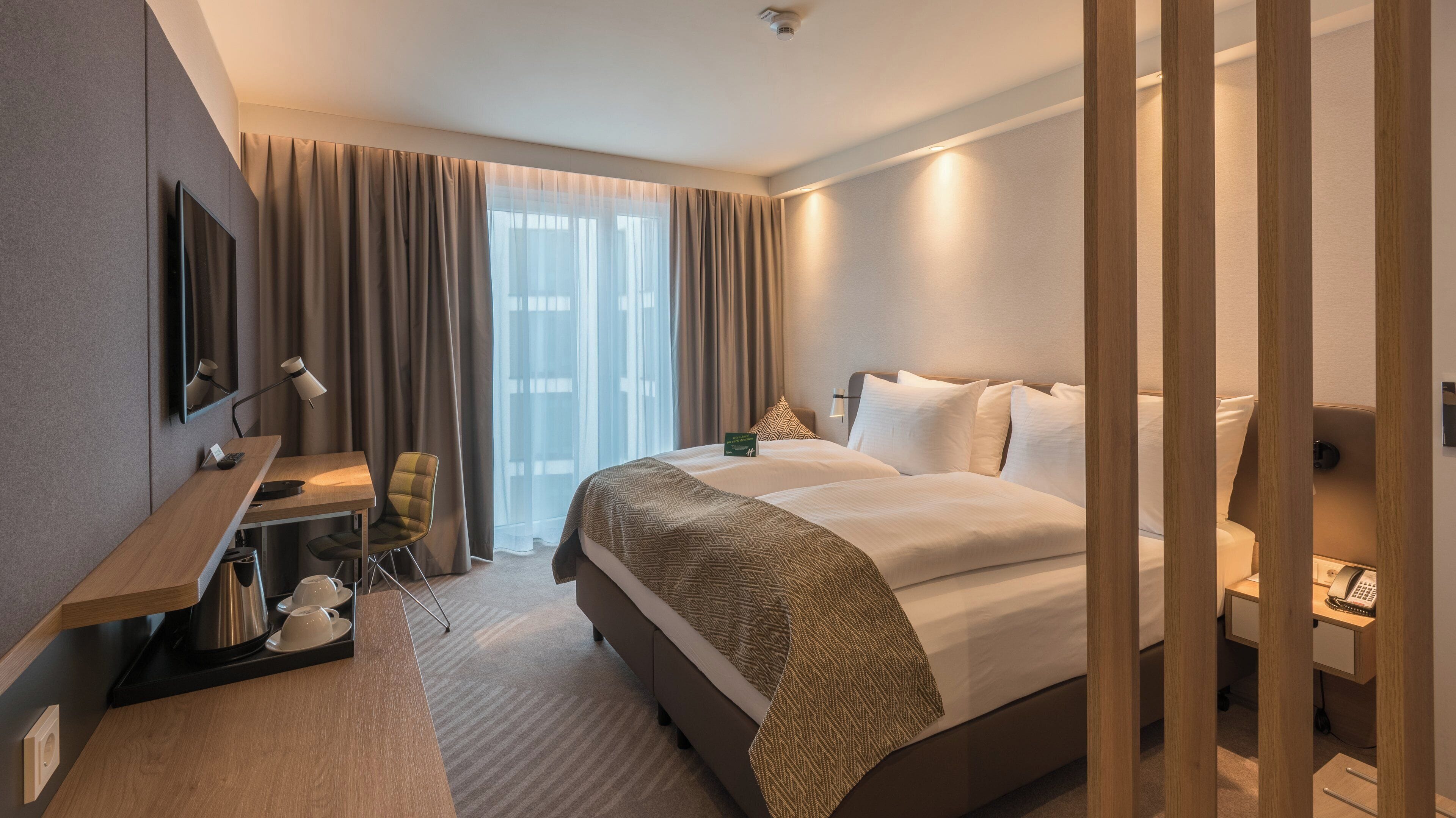 Ein Hotelzimmer mit Bett, Nachttisch, Lampe, Sessel und Schreibtisch mit Telefon.