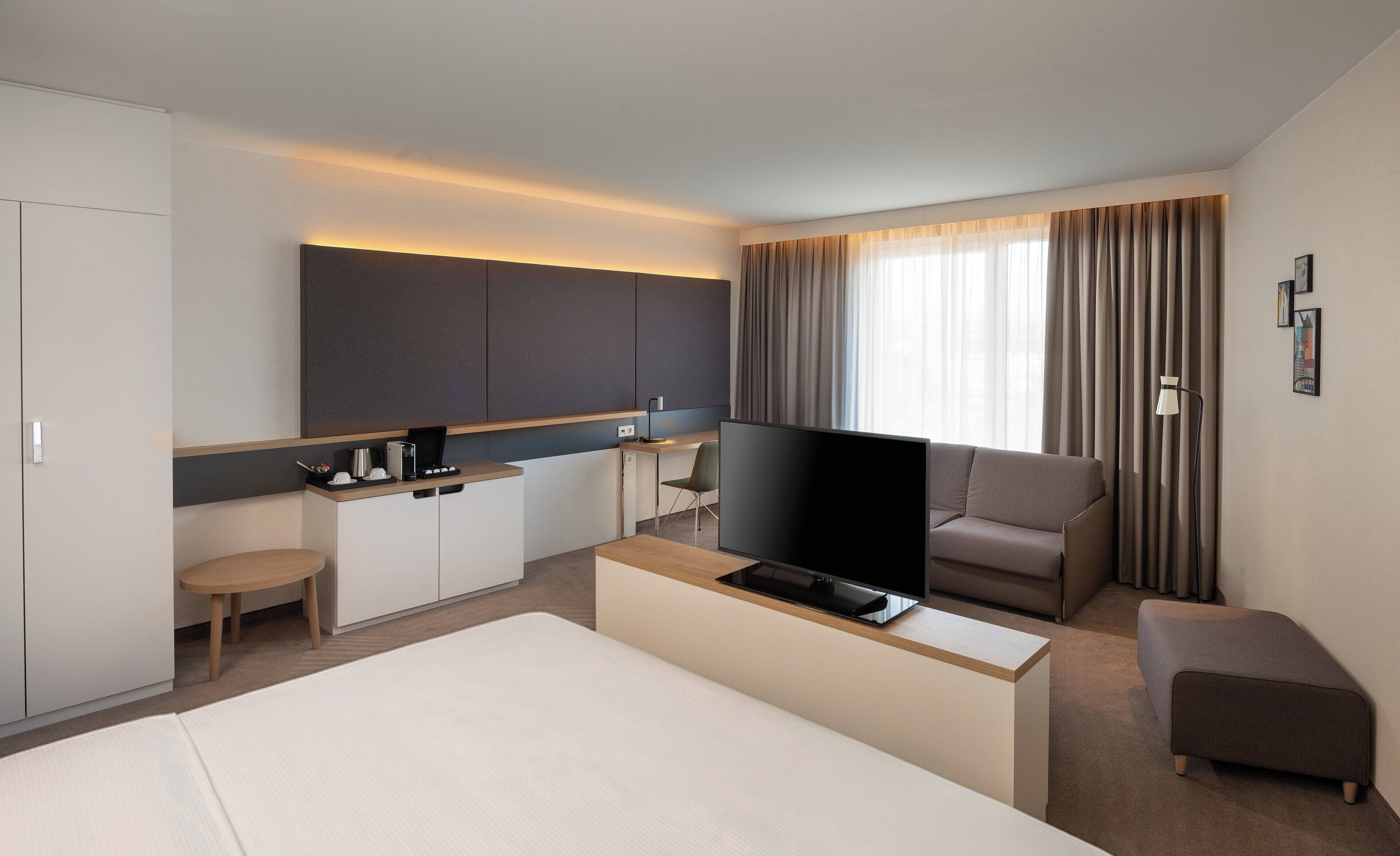 A hotel room with a bed, a TV, a desk with a chair, a kitchenette, and a large window with curtains.
