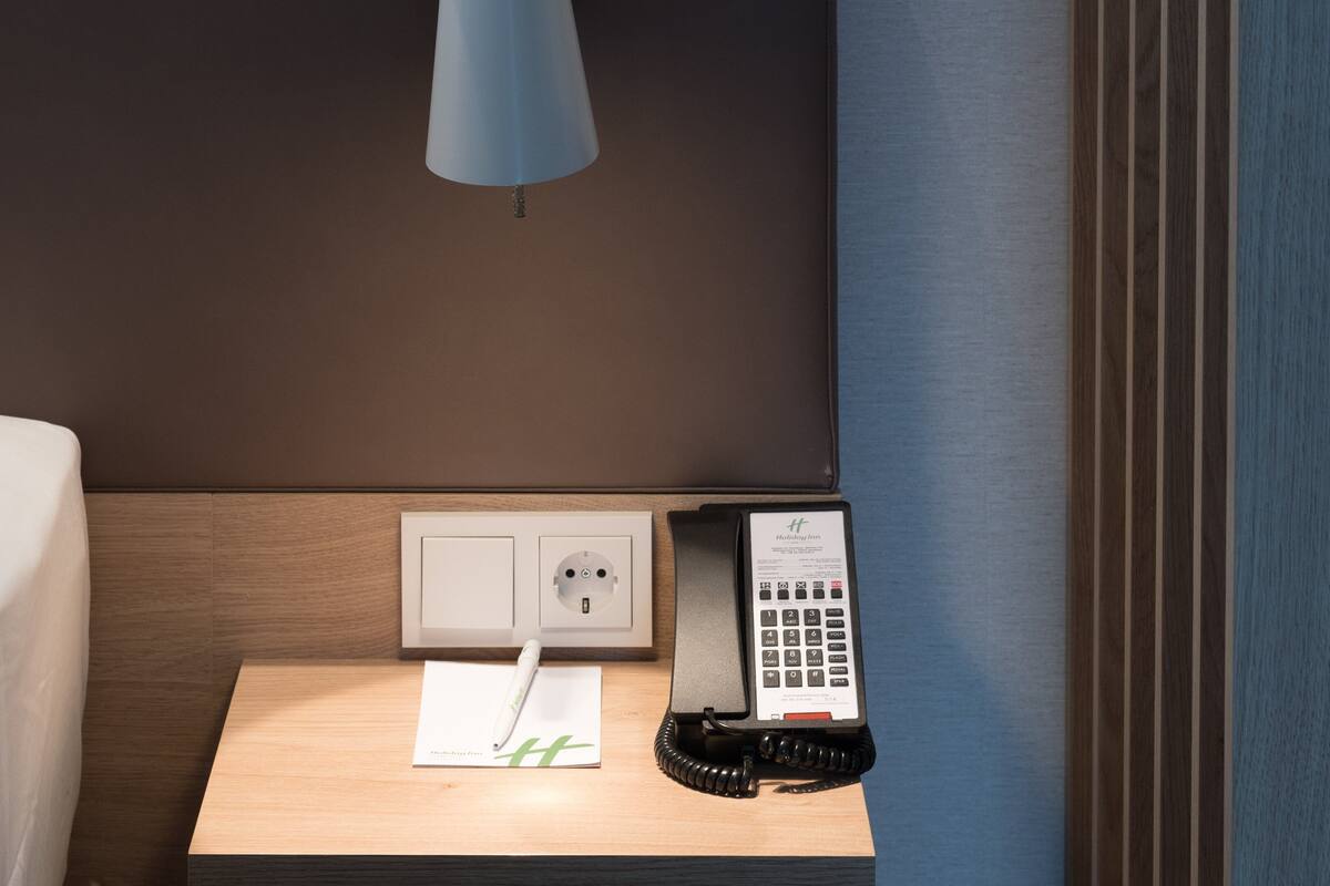 A hotel room with a bed, a bedside table, a lamp, a power outlet, and a telephone.