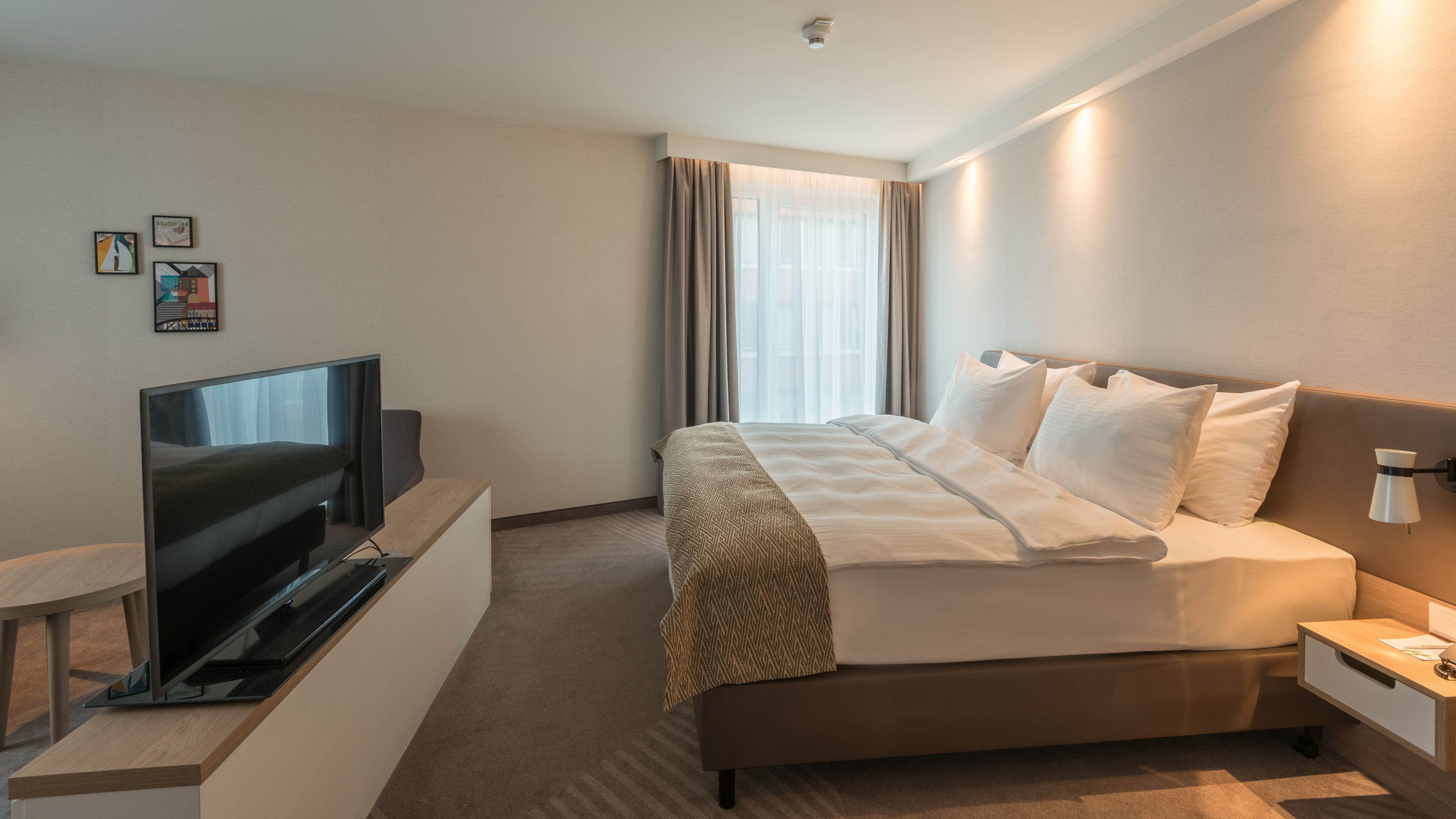 Una habitación de hotel con cama, televisión, mesita de noche y ventana con cortinas.