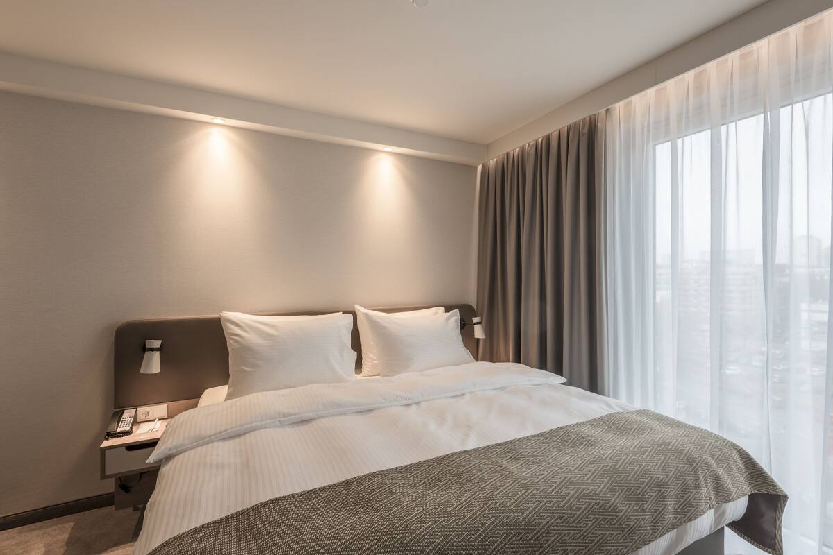 A modern hotel room with a neatly made bed, a bedside telephone, and a window with sheer curtains.