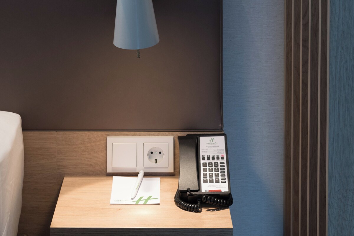 A hotel room with a bed, a bedside table, a lamp, a power outlet, and a telephone.