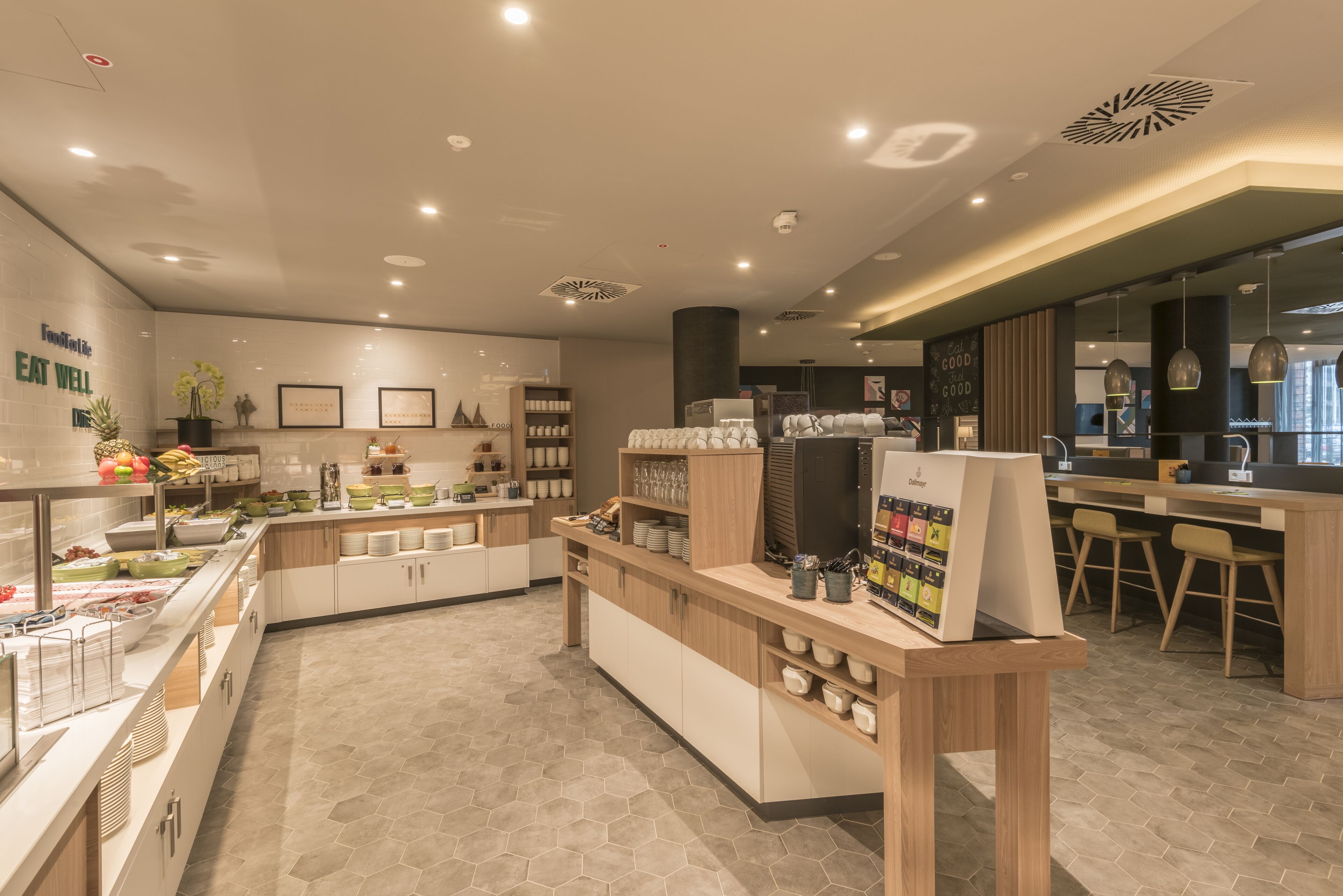 Una cafetería moderna con barra para desayunos, zona de estar y diversas opciones de comida y bebida.