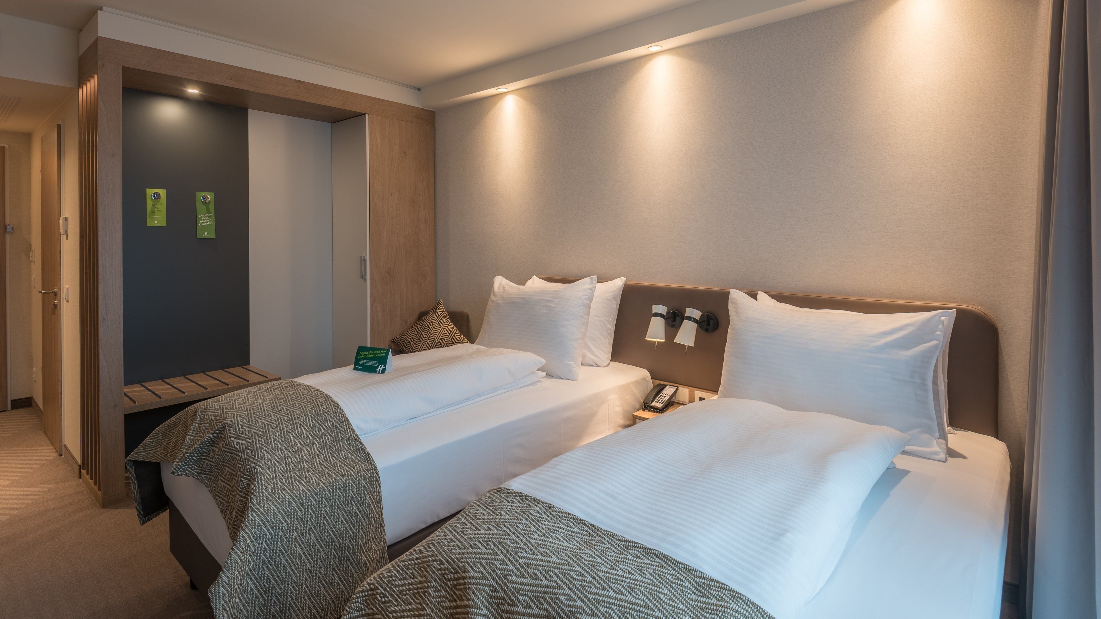A hotel room with two beds, a wooden wardrobe, a bedside table with a lamp, and a phone.