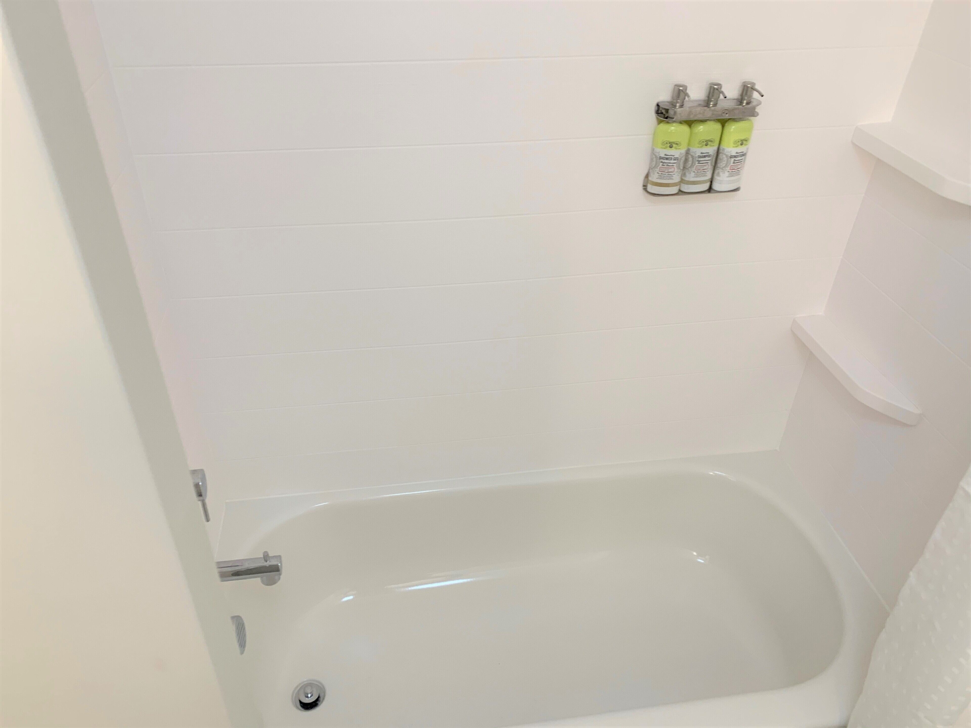 Et badeværelse med et hvidt badekar, bruser og toilet. Tre shampoo flasker hænger på væggen.