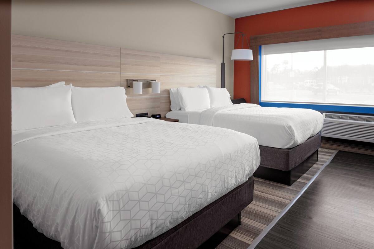 A hotel room with two beds, a large window with a view, a modern lamp, and a wall-mounted air conditioning unit.