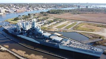 A large naval ship docked at a port with a river and cityscape in the background.