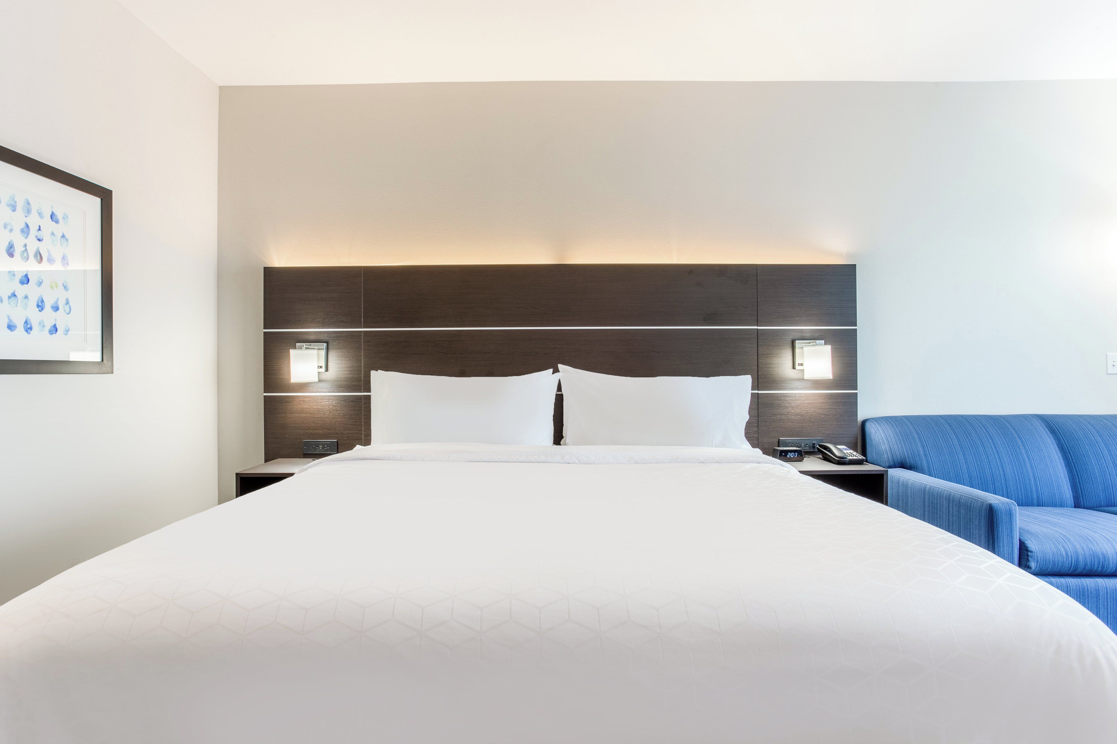 A hotel room with a large bed, two bedside tables, a nightstand, a blue armchair, and a framed picture on the wall.