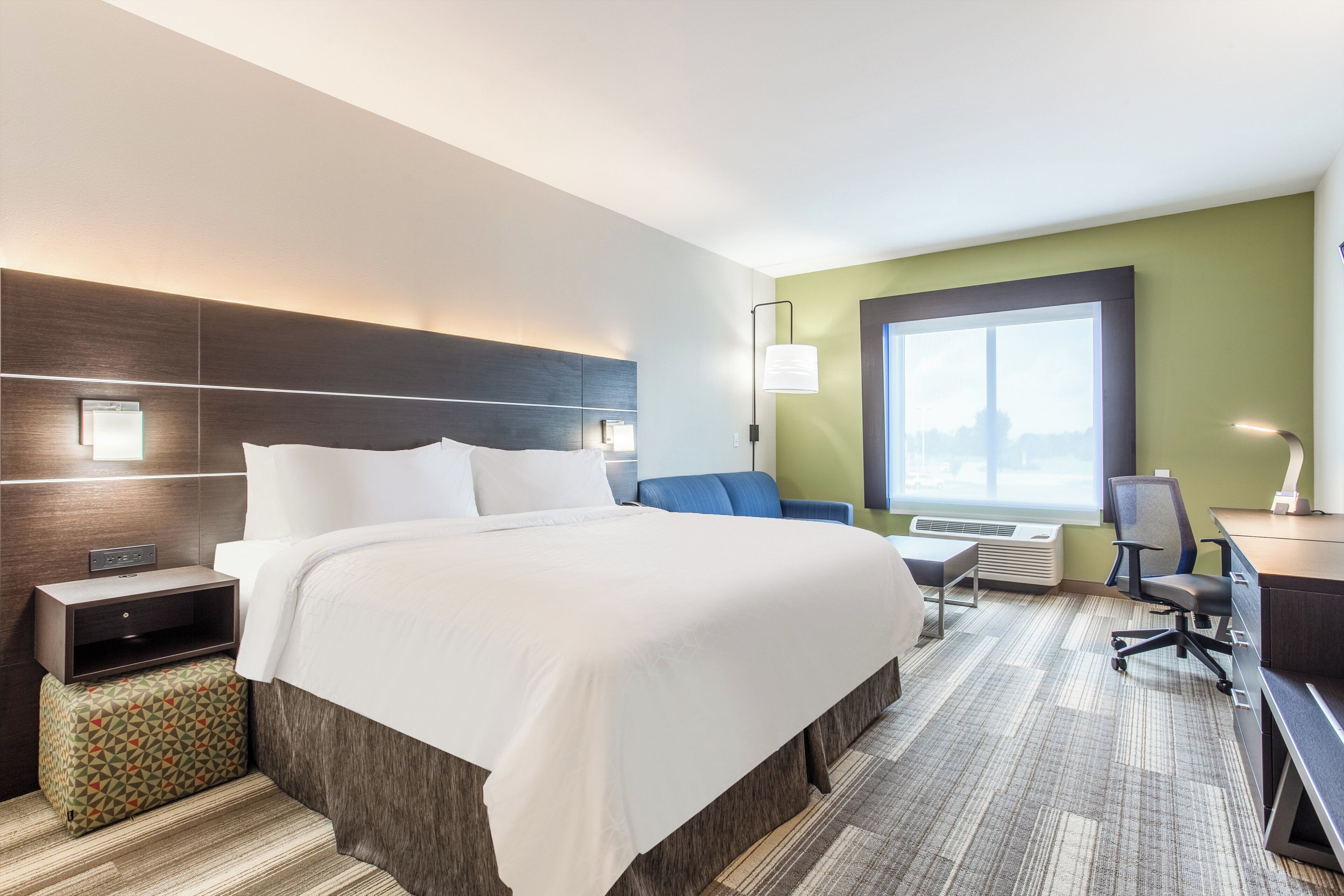 A hotel room with a large bed, a desk, a chair, a nightstand, a lamp, and a window with a view.
