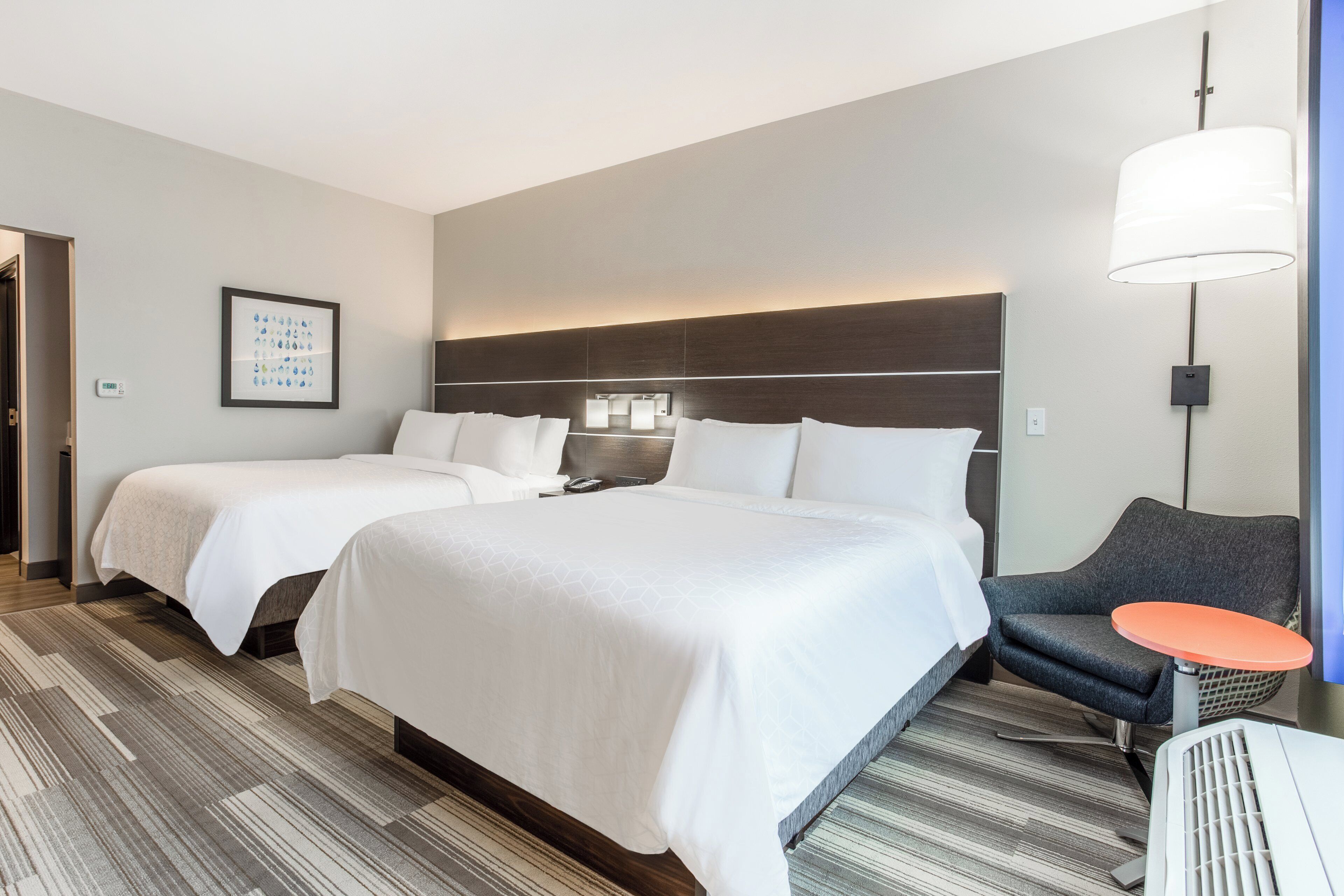 A hotel room with two beds, a desk, a chair, a lamp, and a thermostat on the wall.
