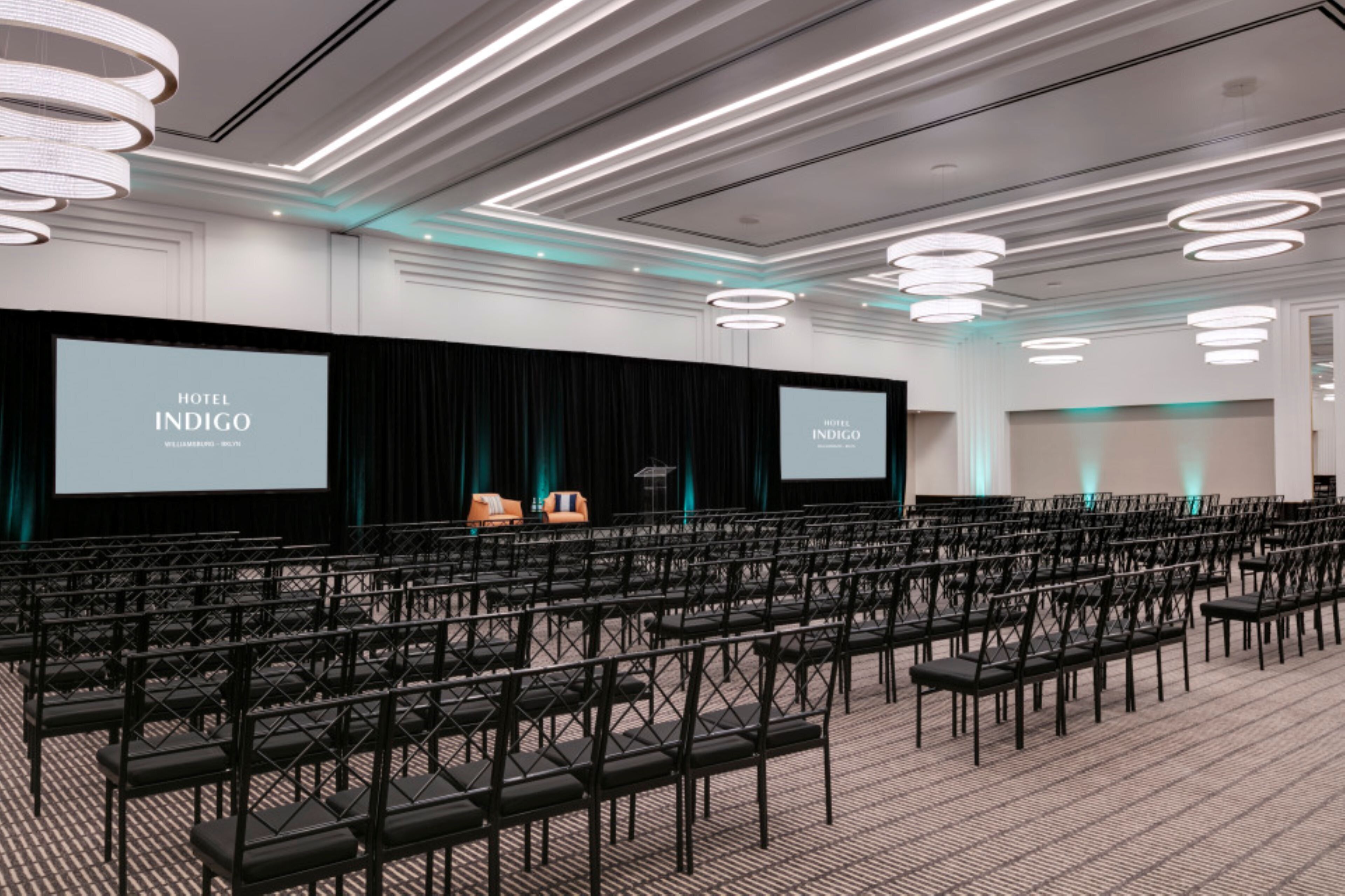A conference room at Hotel Indigo with rows of chairs, a stage with a screen, and modern lighting fixtures.