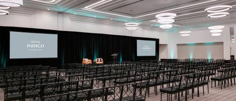 A conference room at Hotel Indigo with rows of chairs, a stage with a screen, and modern lighting fixtures.