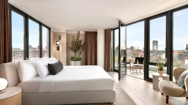 Un dormitorio moderno con una cama grande, una zona de estar con una silla, una planta en maceta y vistas de la ciudad a través de amplias ventanas.