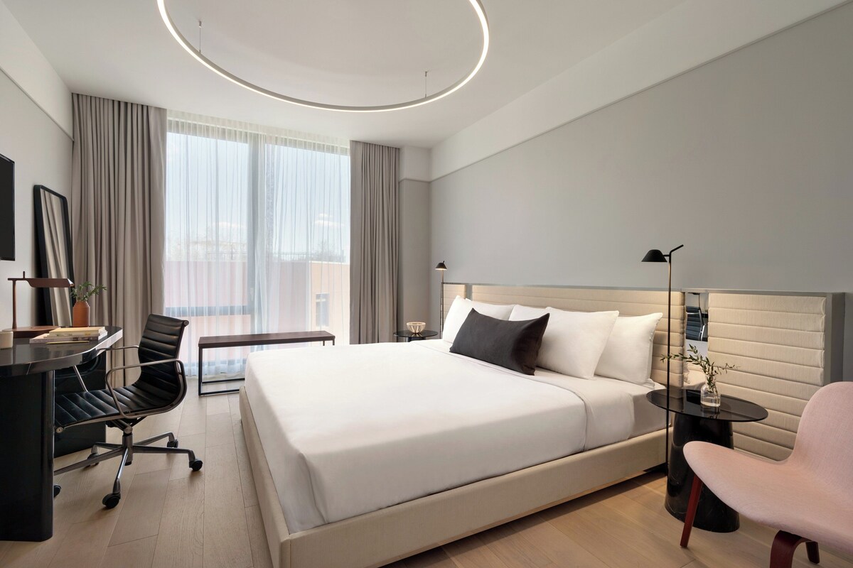 A modern hotel room with a large bed, a desk, a chair, a small table, and a lamp.