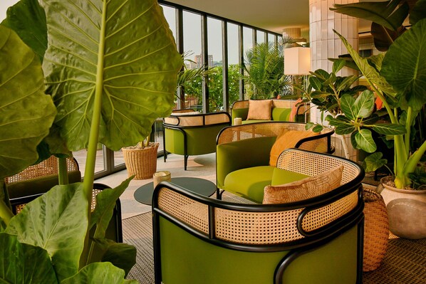 A modern indoor seating area with green upholstered chairs and wicker accents, surrounded by large potted plants.