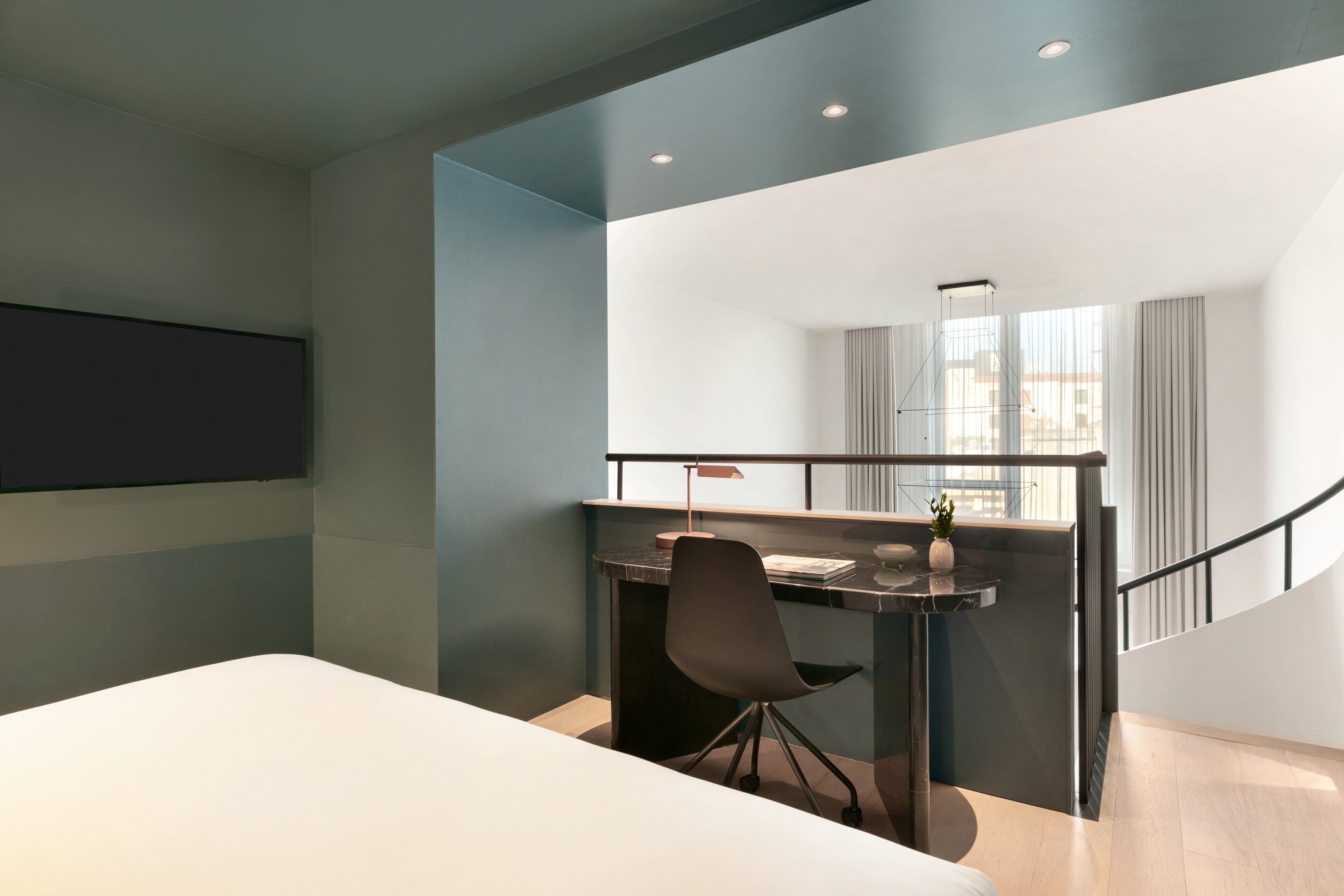 A modern hotel room with a bed, a desk with a chair, a small table, and a window with curtains.