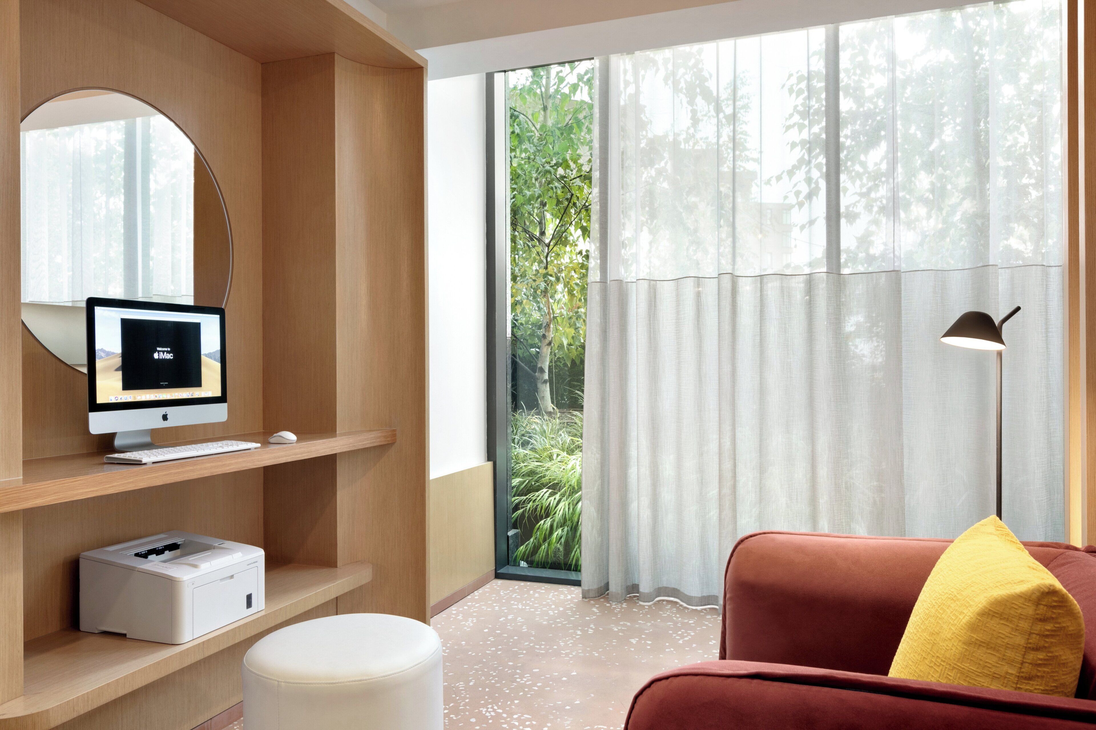 Ein modernes Zimmer mit einem Computer auf einem Holzregal, einem Drucker, einem runden weißen Hocker, einem roten Sofa mit gelbem Kissen und einem großen Fenster mit transparenten Vorhängen.