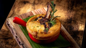 A roasted lamb shank on a red serving dish with herbs, placed on a wooden board with a green leaf underneath.