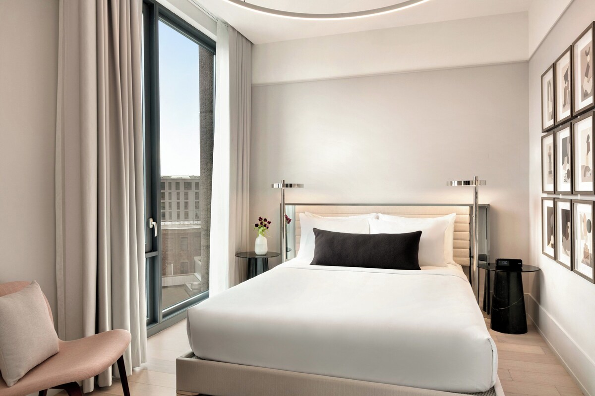 A modern hotel room with a large bed, a chair, a small table, and a window with curtains.
