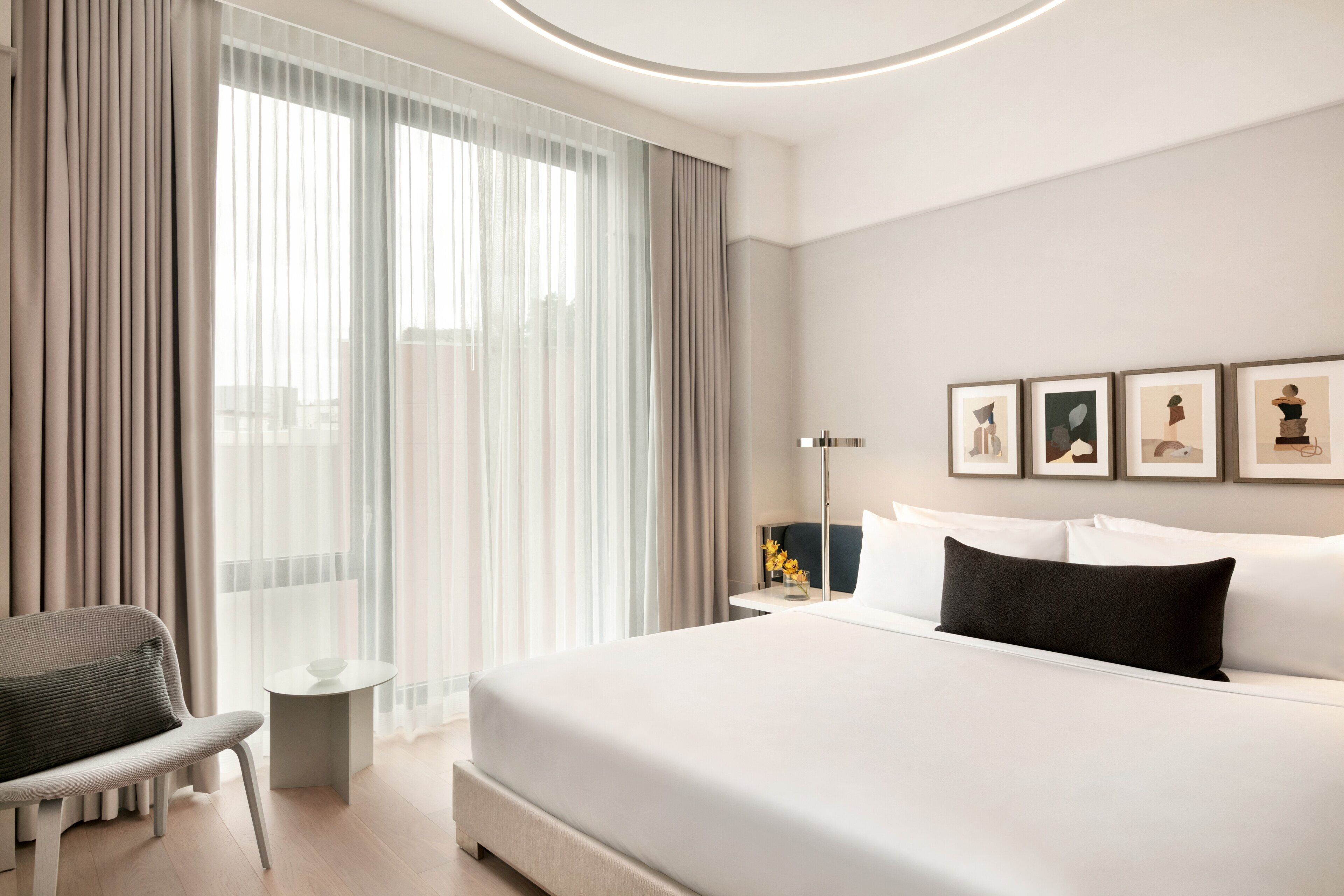 Una habitación de hotel moderna con una cama grande, una silla, una mesita y vistas a la ciudad a través de cortinas translúcidas.