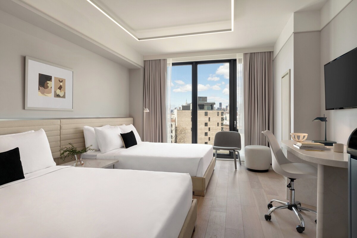 A hotel room with two beds, a desk, a chair, a TV, and a large window with a city view.