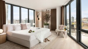 Una habitación de hotel moderna con una cama grande, una zona de estar y vistas de la ciudad a través de amplias ventanas.