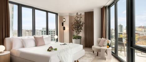 Una habitación de hotel moderna con una cama grande, una zona de estar y vistas de la ciudad a través de amplias ventanas.