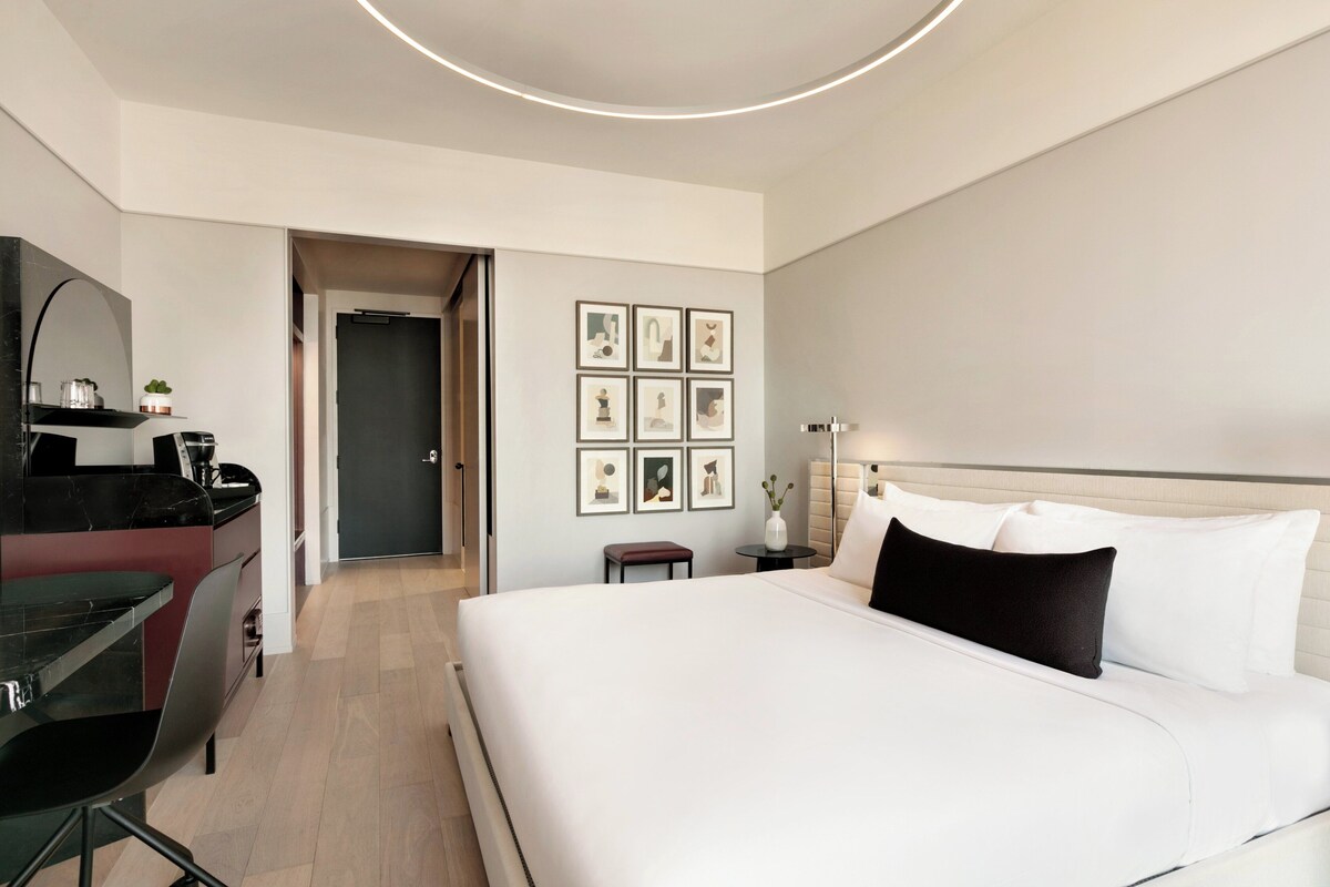A modern hotel room with a large bed, a desk, and a small table. There are framed pictures on the wall and a vase with flowers on the table.