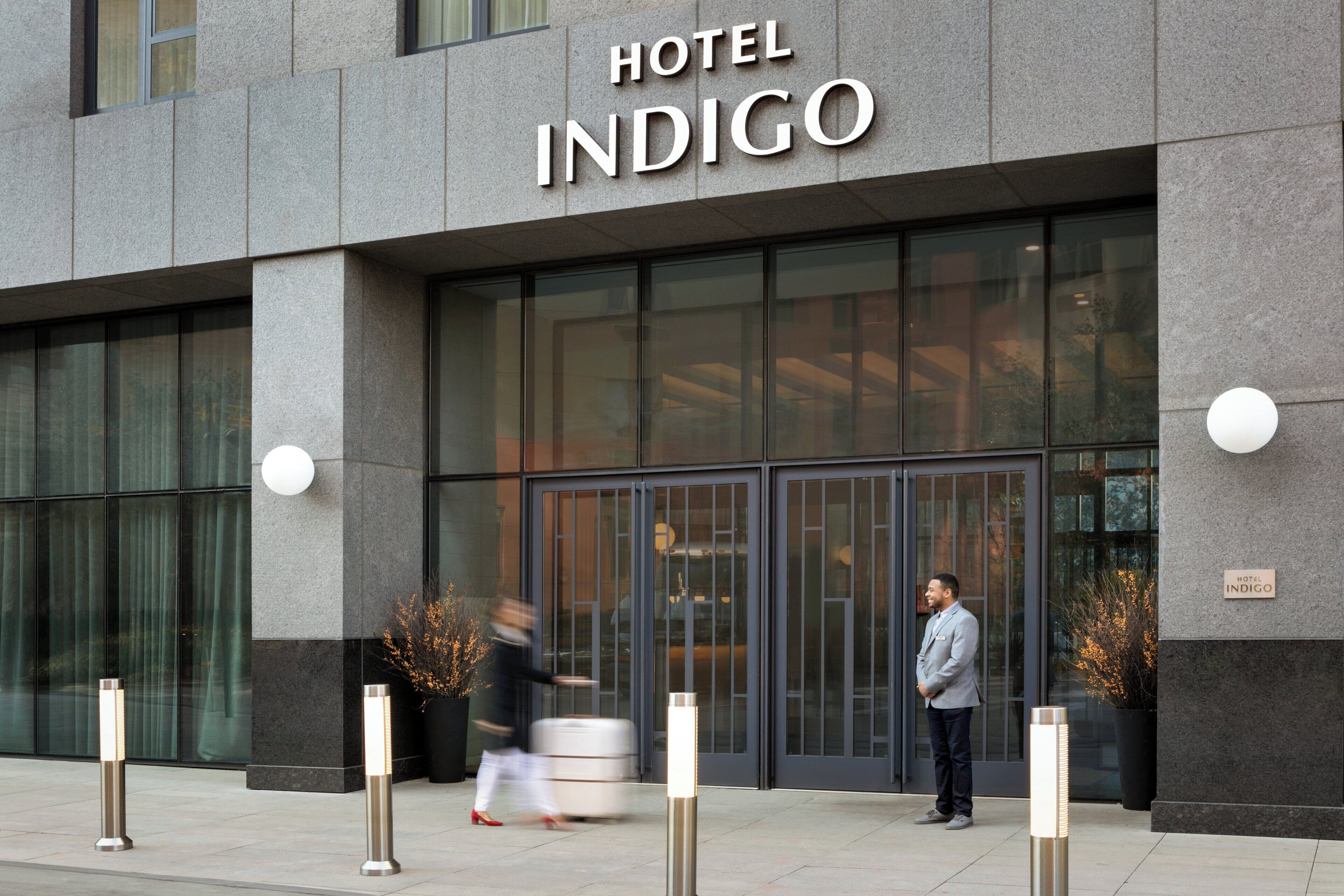 A modern hotel entrance with a large glass facade, a sign reading 'HOTEL INDIGO', and bollards along the sidewalk.