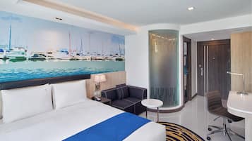 Habitación de hotel con cama, escritorio, silla, una mesita y un cuadro de barcos en la pared.