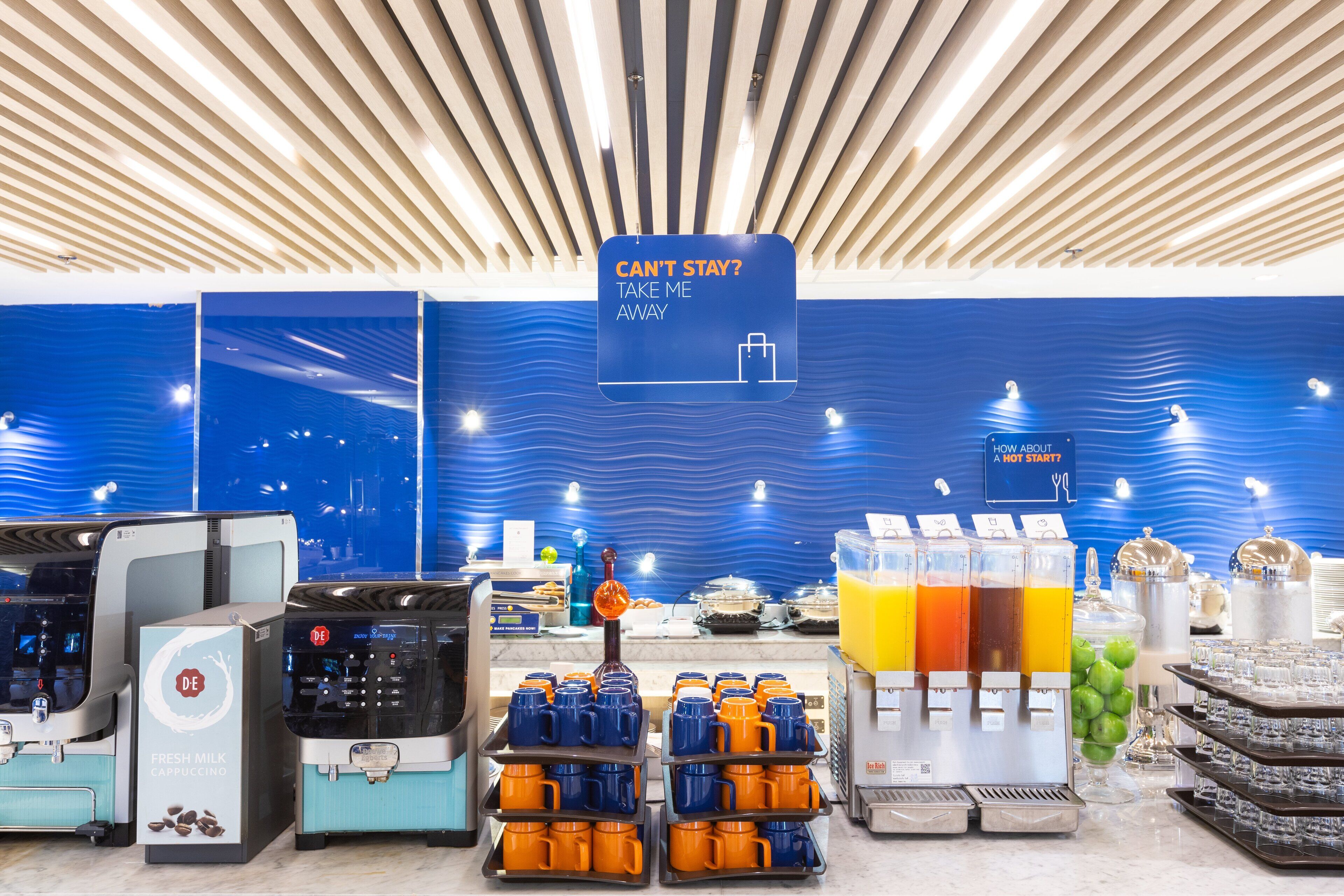 A breakfast buffet area with self-serve coffee machines, a sign offering assistance, and a variety of beverages and food items.