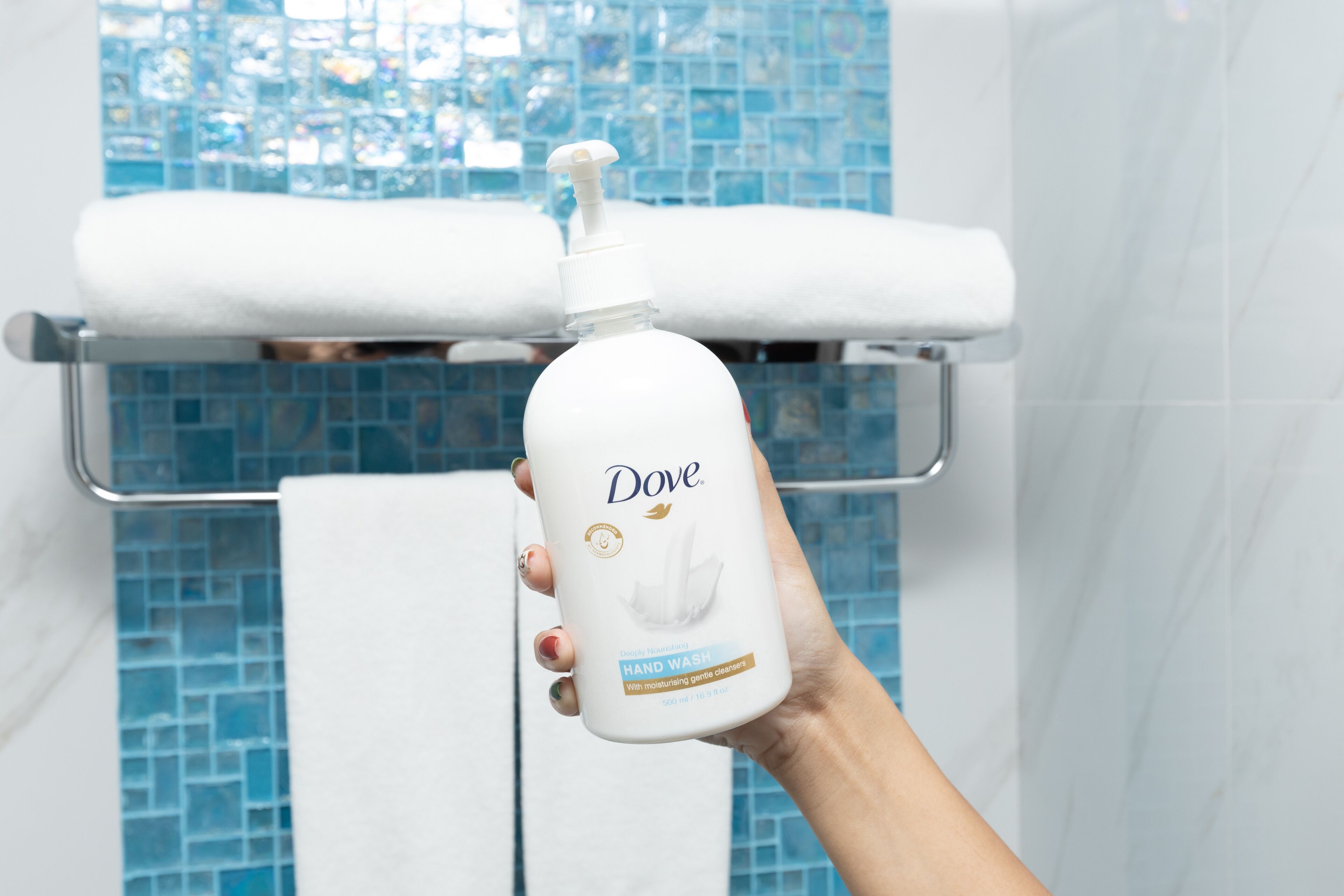 A hand holding a Dove hand wash bottle in a bathroom with a tiled wall and a towel rack.