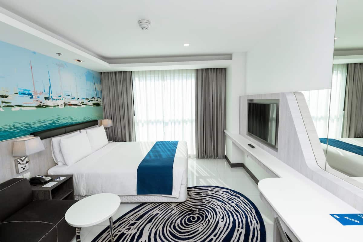Une chambre d’hôtel moderne avec un grand lit, une télévision, un bureau et un tableau représentant des bateaux accroché au mur.