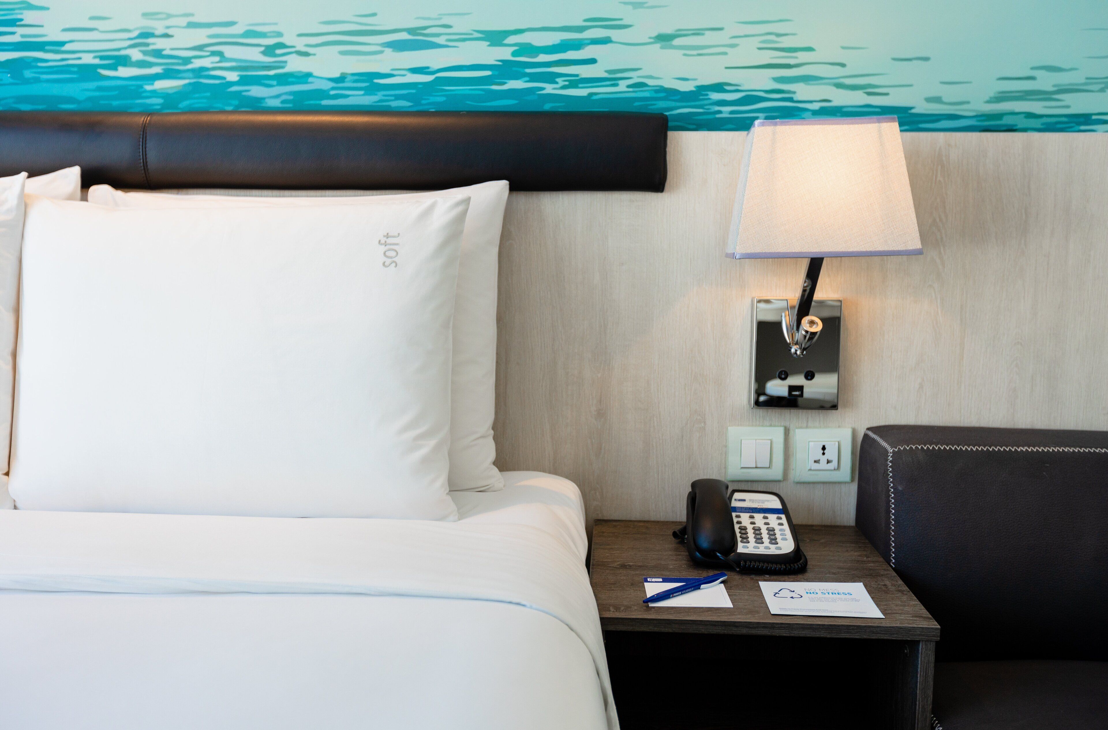 A hotel room with a bed, a nightstand with a lamp, a phone, and a chair.