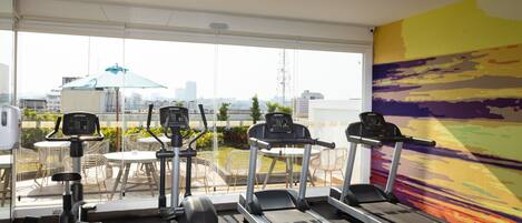 A gym with treadmills, dumbbells, and exercise chairs, overlooking a cityscape.