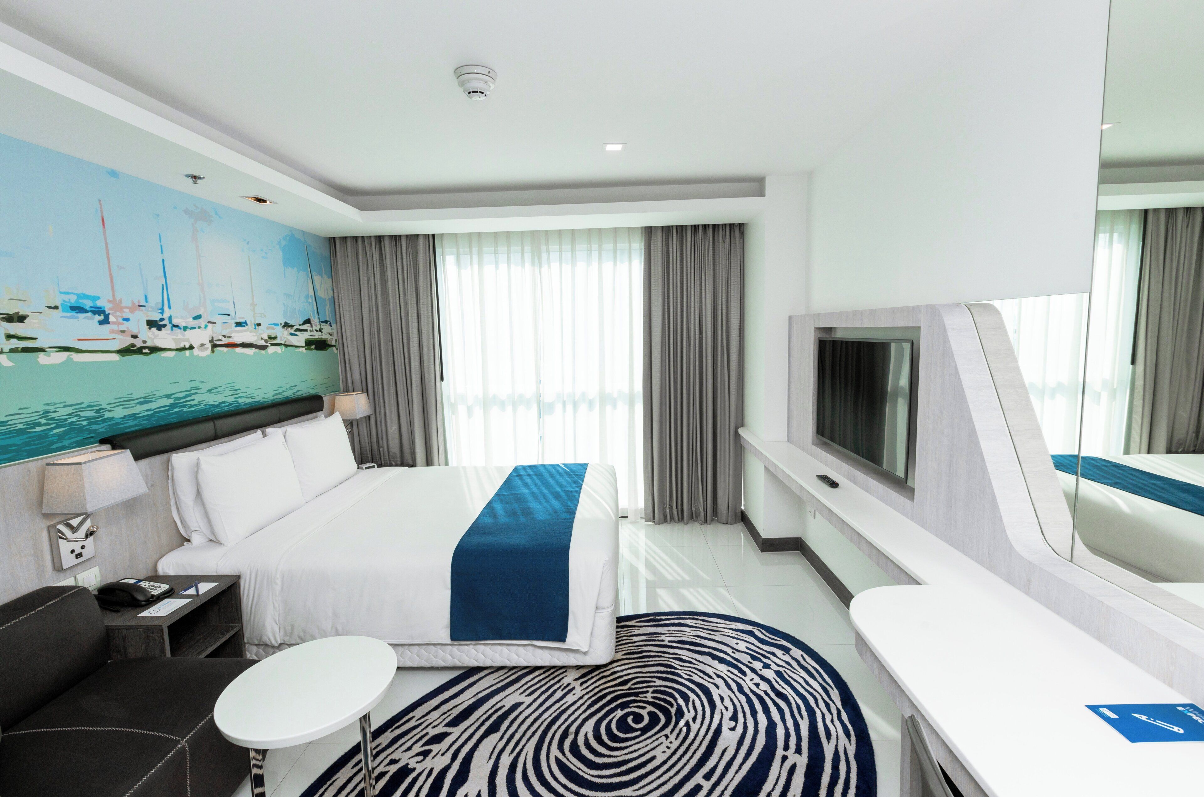 Une chambre d’hôtel moderne avec un grand lit, une télévision, un bureau et un tableau représentant des bateaux accroché au mur.
