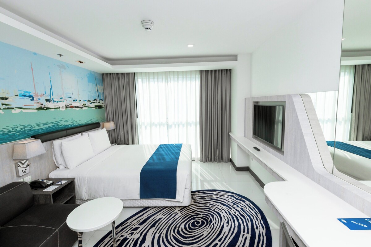 Een moderne hotelkamer met een groot bed, een televisie, een bureau en een schilderij van boten aan de muur.