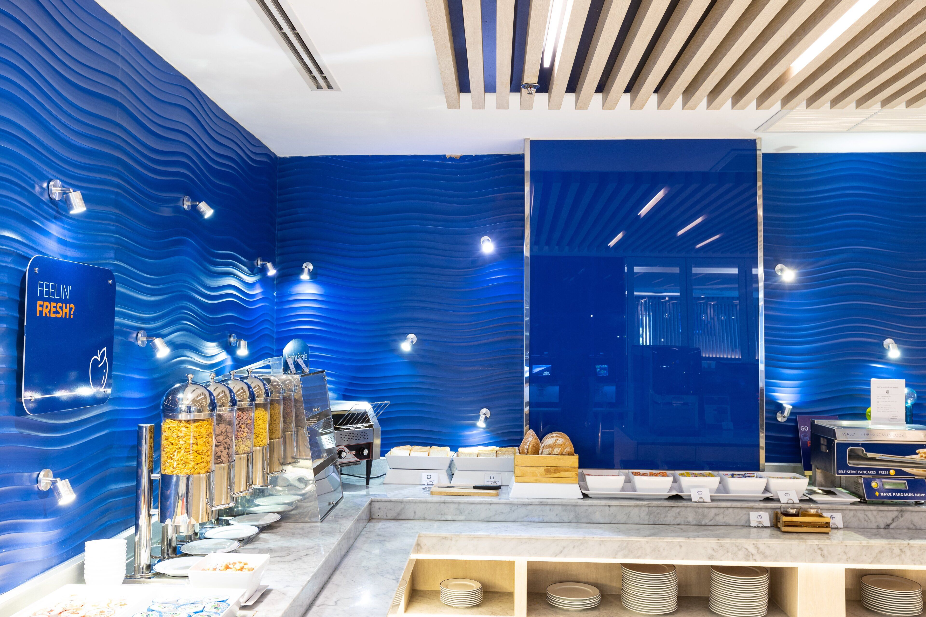 A breakfast buffet with self-serve stations, a large mirror, and a blue wavy wall design.